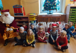 Dzieci siedzą z Mikołajem przy choince.